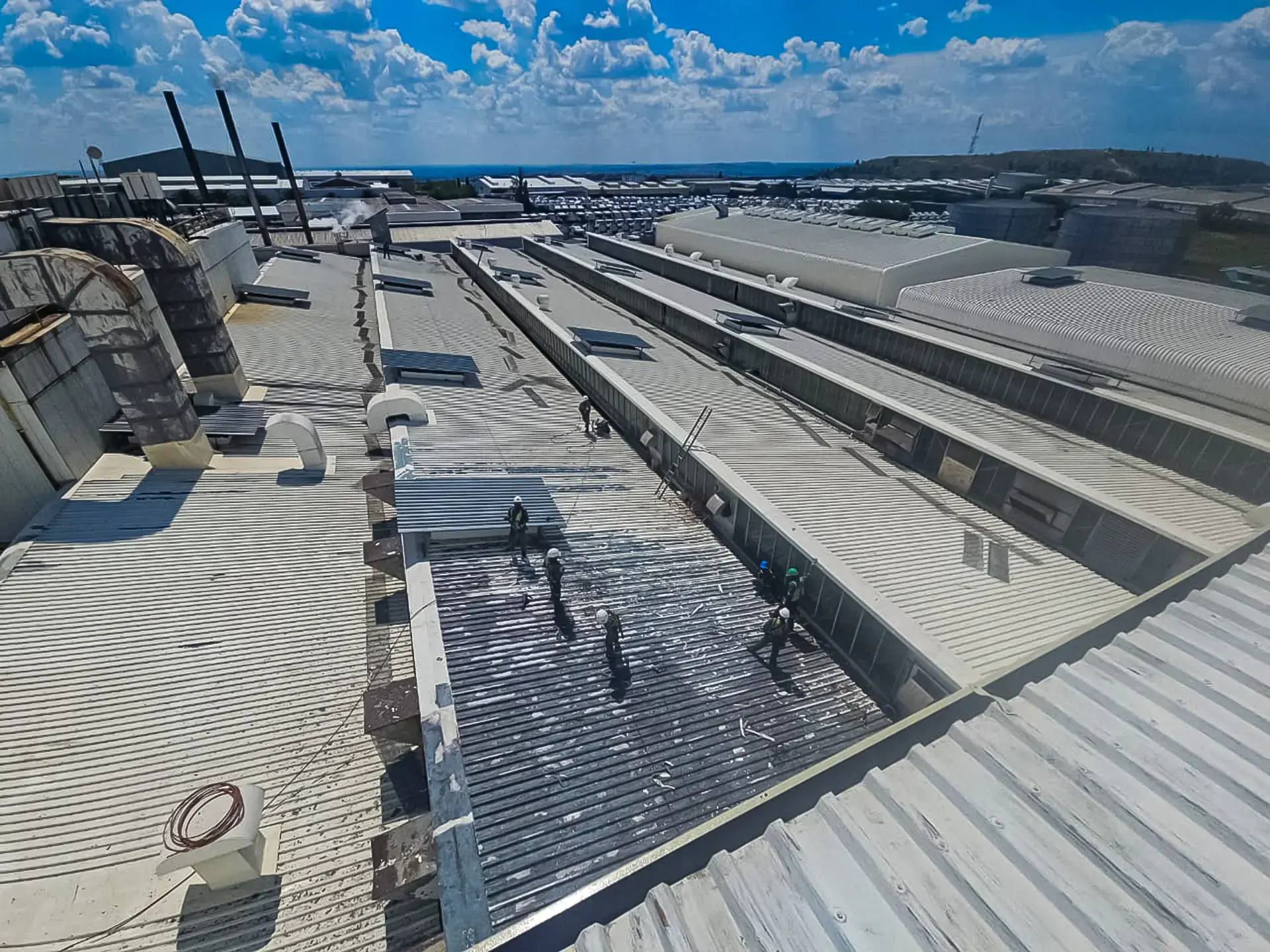 Workers apply new waterproof coating to a large industrial roof under a blue sky, ensuring leak protection for facilities near you.